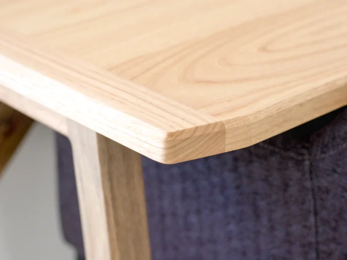 Coin d'une table en bois clair avec des bords arrondis, sur un fond de textile sombre.
